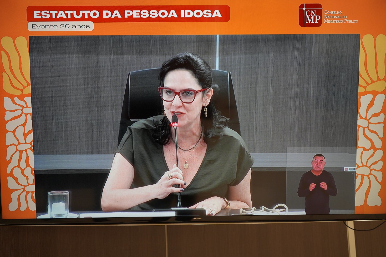 Foto da membra auxiliar Andrea Teixeira, falando enquanto segura um microfone fixo na mesa. A imagem é de uma tela de tv. No canto inferior direito há a imagem de um homem que traduz o que ela diz para Libras. Toda a imagem está inserida na moldura do Seminário, onde se lê no topo Estatuto da Pessoa idosa. Evento 20 anos.